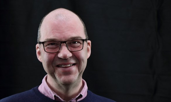 Headshot of Colin Crooks, bald man wearing glasses, navy jumper and pink collared shirt